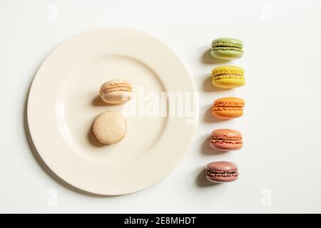 Due macaron leggeri su una piastra luminosa e una fila di macaron di colori diversi su un tavolo bianco, vista dall'alto Foto Stock