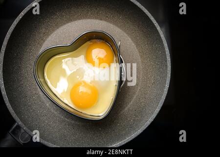 Due uova fritte a forma di cuore su una padella sul fornello nero, preparazione per una romantica colazione valentines giorno, spazio copia, alto angolo vista f Foto Stock