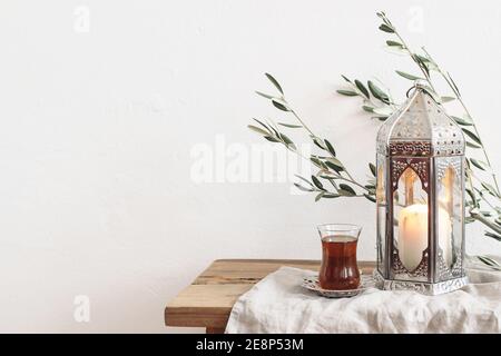 Ramadan Kareem biglietto d'auguri, invito. Lanterna d'argento con candela bruciante. Tè turco in bicchiere sul piatto. Rami di ulivo verde su legno vecchio Foto Stock