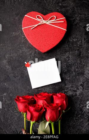 Scatola regalo a forma di cuore rosso, rose, carta messaggio vuota su un tavolo nero grunge. San Valentino sfondo. Foto Stock