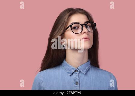 Attraente donna pensiva con capelli lunghi in occhiali e guardando la macchina fotografica, ha un aspetto piacevole, isolato su sfondo rosa. Foto Stock