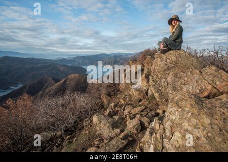 Una donna si siede in cima ad alcune rocce che si affacciano sulla valle e il bacino idrico sotto nella natura selvaggia della California. Foto Stock