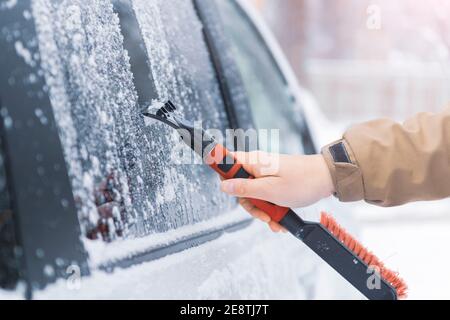 La mano del conducente con raschiatore rimuove il ghiaccio dal parabrezza dell'auto dopo una forte nevicata. Previsioni meteorologiche avverse per la guida. Concetto di sicurezza su strada. Selez Foto Stock