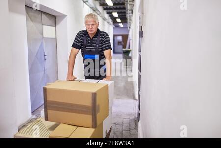Il lavoratore anziano spinge i pacchetti sui carrelli di spinta alle merci in arrivo nel magazzino Foto Stock