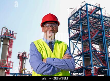 Happy Manager in Red Hardhat, Blue Shirt e Yellow Vest in piedi di fronte alla raffineria di petrolio e gas. Ritratto di Foremen, Supervisore o uomo d'affari Foto Stock