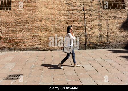 Una donna che cammina mentre parla con un telefono cellulare Sulla vecchia parete sotto il sole nella città di Bologna Italia Foto Stock