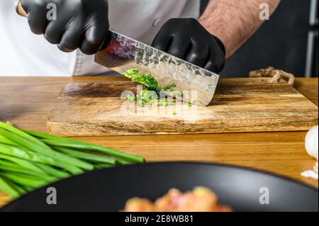 Uno chef in guanti neri sta affettando cipolle verdi fresche su un tagliere di legno. Foto Stock