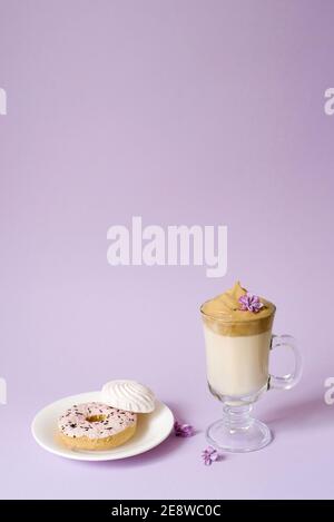 Bella dalgona bere un caffè di schiuma in una tazza trasparente e fiori di lilla su uno sfondo viola. Dolci su un piatto: Donat e tenero marshmal Foto Stock