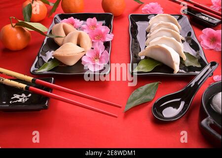 Cena orientale festiva. Cinese nuovo anno cibo e bevande ancora vita. Gnocchi al vapore, palle di riso Tangyuan, biscotti fortune su piatto nero e sfondo rosso. Foto di alta qualità. Sopra Foto Stock