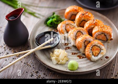 Panini al sushi maki al forno con salmone, granchio e salsa piccante su un piatto con chopsticks, salsa di soia, wasabi e zenzero. Cibo tradizionale giapponese a base di pesce Foto Stock
