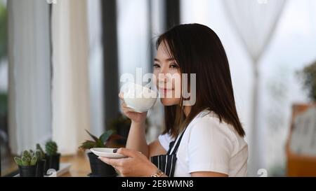 Vista laterale della barista asiatica che tiene una tazza di caffè e puzzava nella caffetteria. Foto Stock