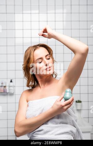 donna in asciugamano bianco che solleva la mano e applica il rotolo deodorante in bagno Foto Stock