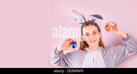 Ragazza in orecchie conigliate di coniglio sulla testa con uova colorate su sfondo rosa. Allegro sorridente bambino felice. Banner delle vacanze di Pasqua Foto Stock