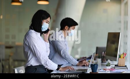 Vista laterale di un dipendente asiatico in ufficio, donna che indossa una maschera protettiva, lavora in un nuovo ufficio normale con il suo collega. Foto Stock