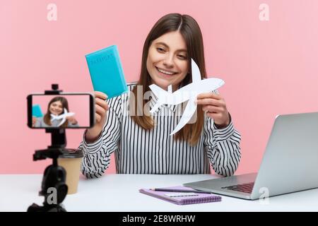 Blogger donna positivo che mostra passaporto e aereo di carta a fotocamera smartphone, vantare circa il tour, andare in vacanza. Studio al coperto isolato Foto Stock