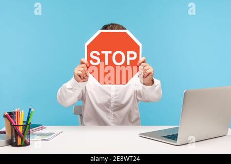 Uomo lavoratore in camicia bianca nascondendo il volto dietro il cartello rosso stop traffico, seduto sul posto di lavoro con il laptop, simbolo di restrizione di divieto. Interno Foto Stock