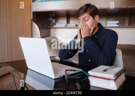 Stressato uomo d'affari malsano stanco, overwork giovane uomo che indossa occhiali che soffrono di forte mal di testa, seduto alla scrivania con computer portatile Foto Stock
