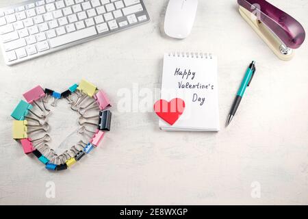 Le parole Happy Valentines giorno in una nota e cuore rosso sul desktop con clip cardio colorate. San Valentino in ufficio. Vista dall'alto Foto Stock