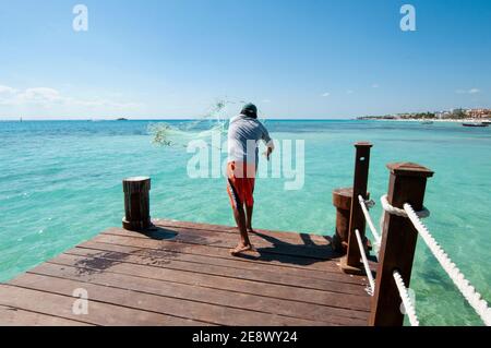 Un pescatore da dietro getta la sua rete da un molo di legno; sullo sfondo il Mar dei Caraibi e la costa di Playa del Carmen, Messico Foto Stock