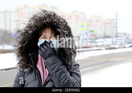 Protezione da coronavirus, donna in maschera medica e cappuccio in pelliccia in piedi sulla strada invernale durante la neve. Concetto di malattia, febbre, raffreddore e influenza Foto Stock