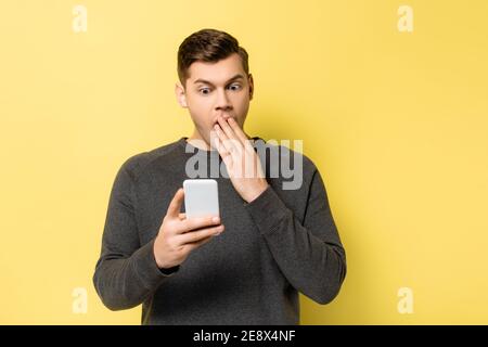 Uomo stupito che copre la bocca mentre tiene lo smartphone su sfondo giallo Foto Stock