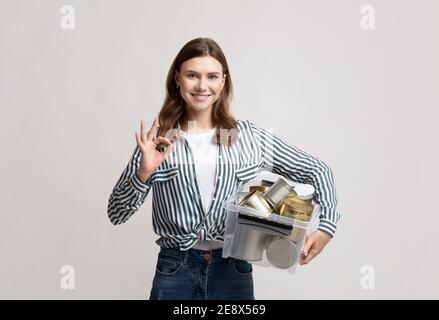 Riciclaggio dei metalli. Contenitore positivo Lady Holding con lattine di stagno, mostrando OK Gesture Foto Stock