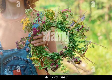 Donna in tuta denim tiene una corona di fiori di foresta. Primo piano. Sfondo della foresta Foto Stock