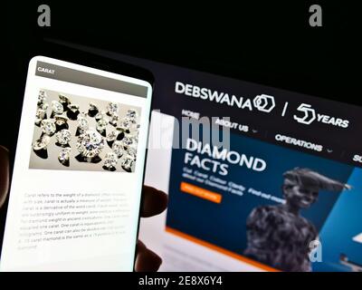 Persona che detiene smartphone con il sito web della società mineraria Debswana Diamond Company Limited davanti al logo. Mettere a fuoco nella parte superiore centrale dello schermo del telefono. Foto Stock