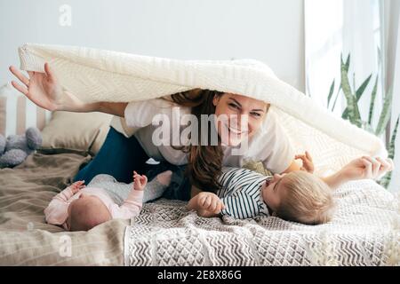 La mamma gioca con i bambini in camera da letto. La mamma copre due bambini con una coperta e ride. Foto Stock