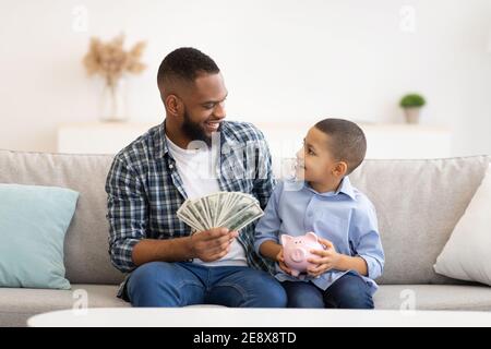 Il padre nero e suo figlio che mette i soldi in Piggybank all'interno Foto Stock