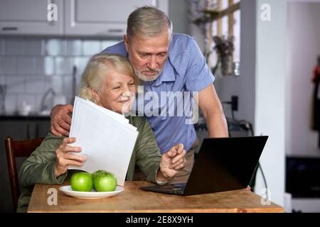 coppie anziane che controllano le finanze a casa usando il laptop, discutono insieme il preventivo di pianificazione, usando i servizi e la calcolatrice di banca in linea, tenendo il documento Foto Stock