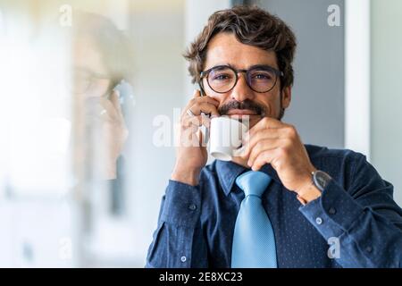 Uomo d'affari che indossa occhiali, sorridendo mentre si parla al telefono e bevendo caffè. Felice ritratto maschile di successo in ufficio presso la finestra Foto Stock