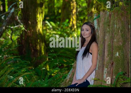 Una giovane donna con lunghi capelli marroni si appoggia contro un albero in un ambiente forestale. Foto Stock