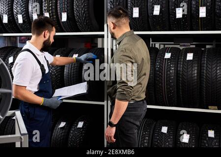 il venditore offre nuove ruote in gomma, pneumatici, cerchioni per l'auto. cliente maschile è venuto a fare l'acquisto nel negozio di servizio auto. meccanico auto mostrando l'assortimento Foto Stock