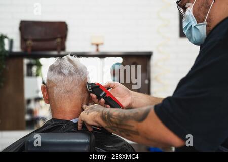 Parrucchiere maschile taglio capelli per hipster senior client mentre si indossa Maschera chirurgica facciale - giovane hairstylist che lavora in barbershop durante corona virus Foto Stock