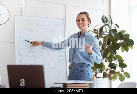 Giovane insegnante gioioso in piedi vicino a lavagna, spiegando la grammatica, dando lezioni online agli studenti sul laptop Foto Stock