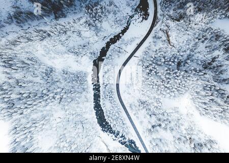 Strada tortuosa che costeggia un fiume ghiacciato nella foresta invernale coperta di neve, vista aerea, Svizzera Foto Stock