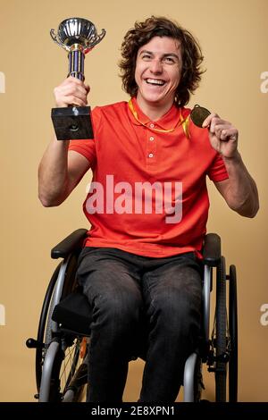 sportivo per disabili paralimpico in sedia a rotelle che tiene gobblet campione e oro medaglie isolate su sfondo beige Foto Stock