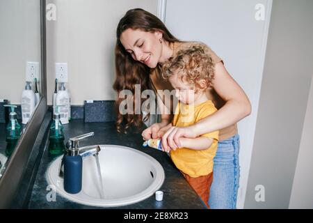 Giovane madre caucasica che aiuta il bimbo a spazzolare i denti in bagno a casa. Igiene sanitaria e routine mattutina per bambini. Mamma che aiuta a supportare te Foto Stock