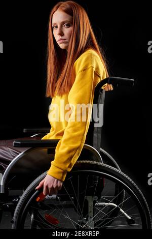 triste donna disabile in una sedia a rotelle, guardando la macchina fotografica depressa. rosso femmina in giallo camicia casual si siede isolato su sfondo nero. salute e p Foto Stock