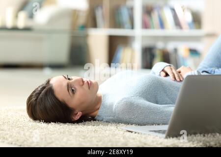 Una donna penosa accanto a un computer portatile si guarda via sdraiata sul pavimento a casa Foto Stock