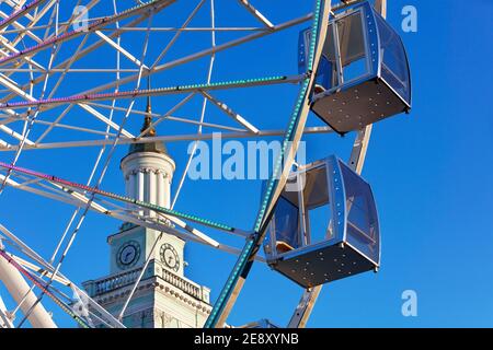 Una bella e panoramica vista delle cabine vuote della ruota panoramica contro il cielo blu e la guglia della vecchia torre dell'orologio nel pomeriggio. Foto Stock