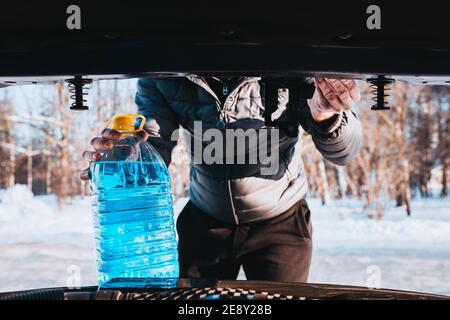 Un uomo ha aperto il cofano e versa una rondella antigelo liquido nel serbatoio - un tergicristallo in inverno Foto Stock