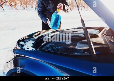 Un uomo ha aperto il cofano e versa una rondella antigelo liquido nel serbatoio - un tergicristallo in inverno Foto Stock