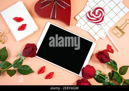 Desktop Happy Valentine's Day con dispositivo tablet, con rose rosse di San Valentino, regalo a forma di cuore e cioccolatini kissess rossetto su un corallo moderno Foto Stock