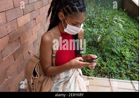 Giovane afrolatina donna in maschera con il suo smartphone in strada, Colombia Foto Stock