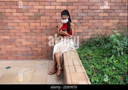 Giovane afrolatina donna in maschera con il suo smartphone in strada, Colombia Foto Stock