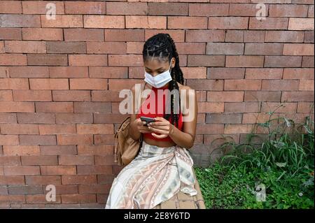 Giovane afrolatina donna in maschera con il suo smartphone in strada, Colombia Foto Stock