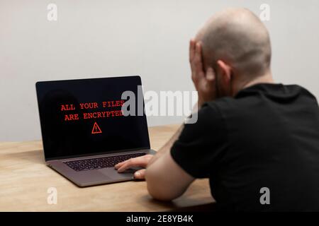 Testo rosso che indica ‘tutti i file sono crittografati’ e un segnale di avviso sullo schermo del computer. Un uomo disperato che guarda il suo laptop. Ransomware, cybercrimine Foto Stock