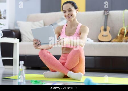 Donna in abbigliamento sportivo seduta sul tappetino ginnico e reggendo la tavoletta nelle mani Foto Stock